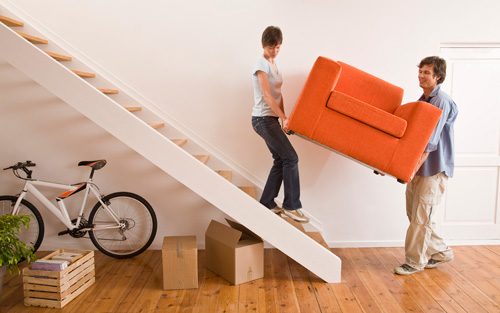 Zwei Personen tragen einen Sessel eine Treppe hinauf, daneben stehen Umzugskartons